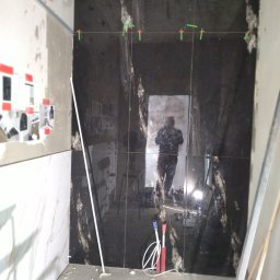 Usługi Remontowo Budowlane - Układanie ciemnych, marmurkowych płytek na ścianie w łazience, widoczne krzyżyki dystansowe, odbicie glazurnika w płytkach, częściowo wykończona ściana z jasnymi płytkami.