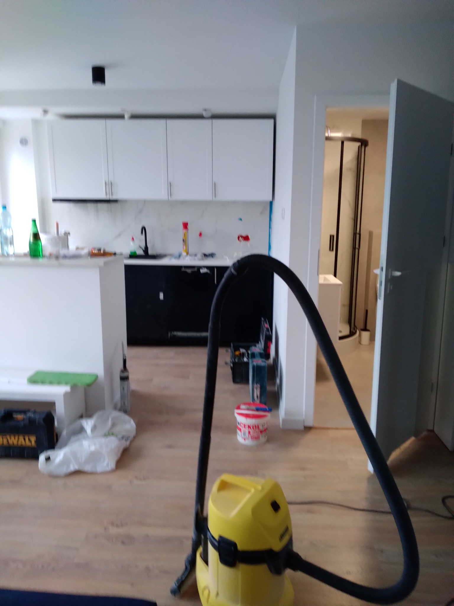 Wnętrze mieszkania w trakcie remontu: widoczny odkurzacz przemysłowy Karcher w kolorze żółtym na parkiecie, w tle kuchnia z białymi szafkami i czarnym blatem oraz otwarta łazienka z kabiną...