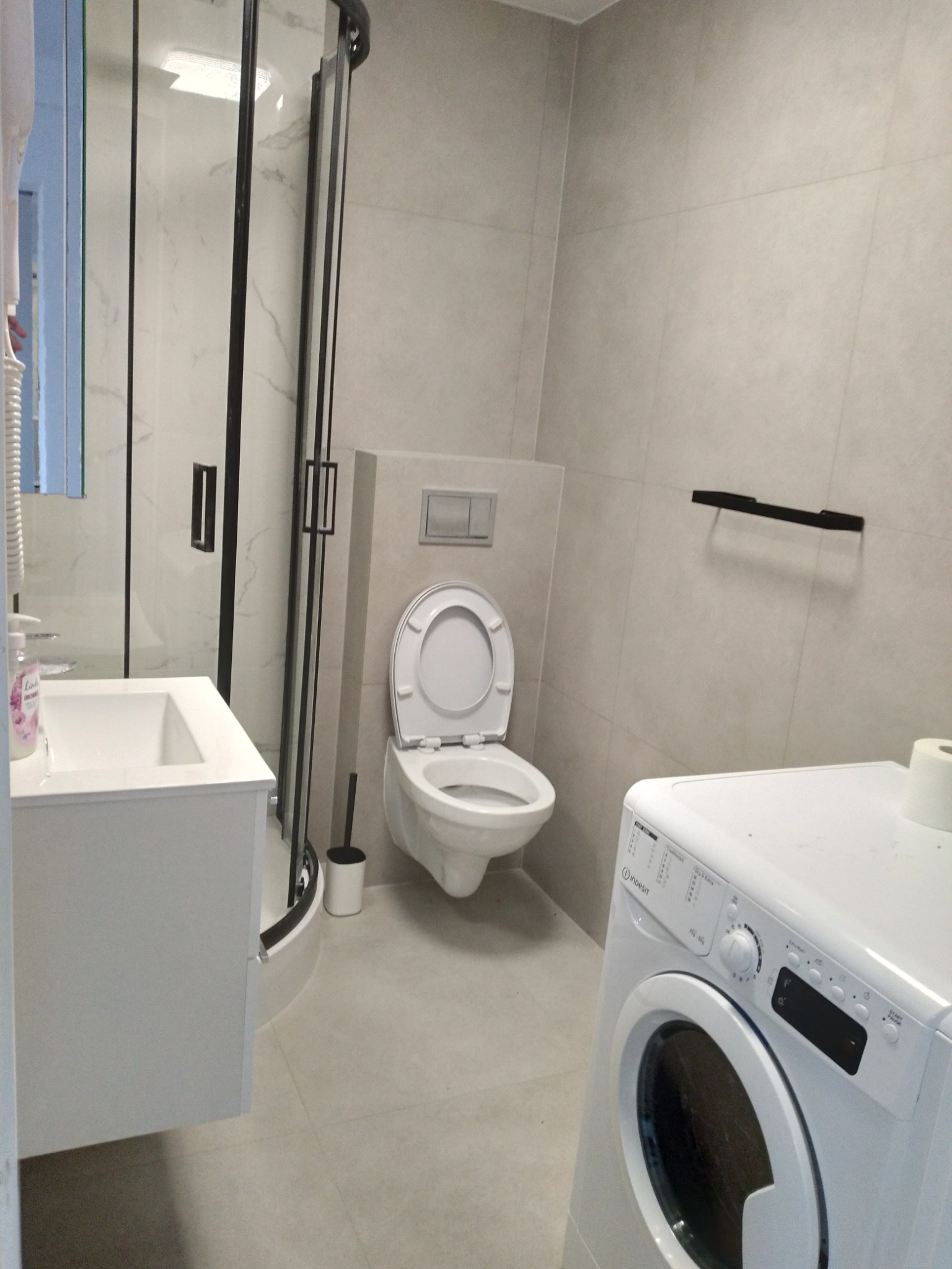 Jasna łazienka z kabiną prysznicową z czarnymi ramami, umywalką z szafką, toaletą i pralką Indesit.