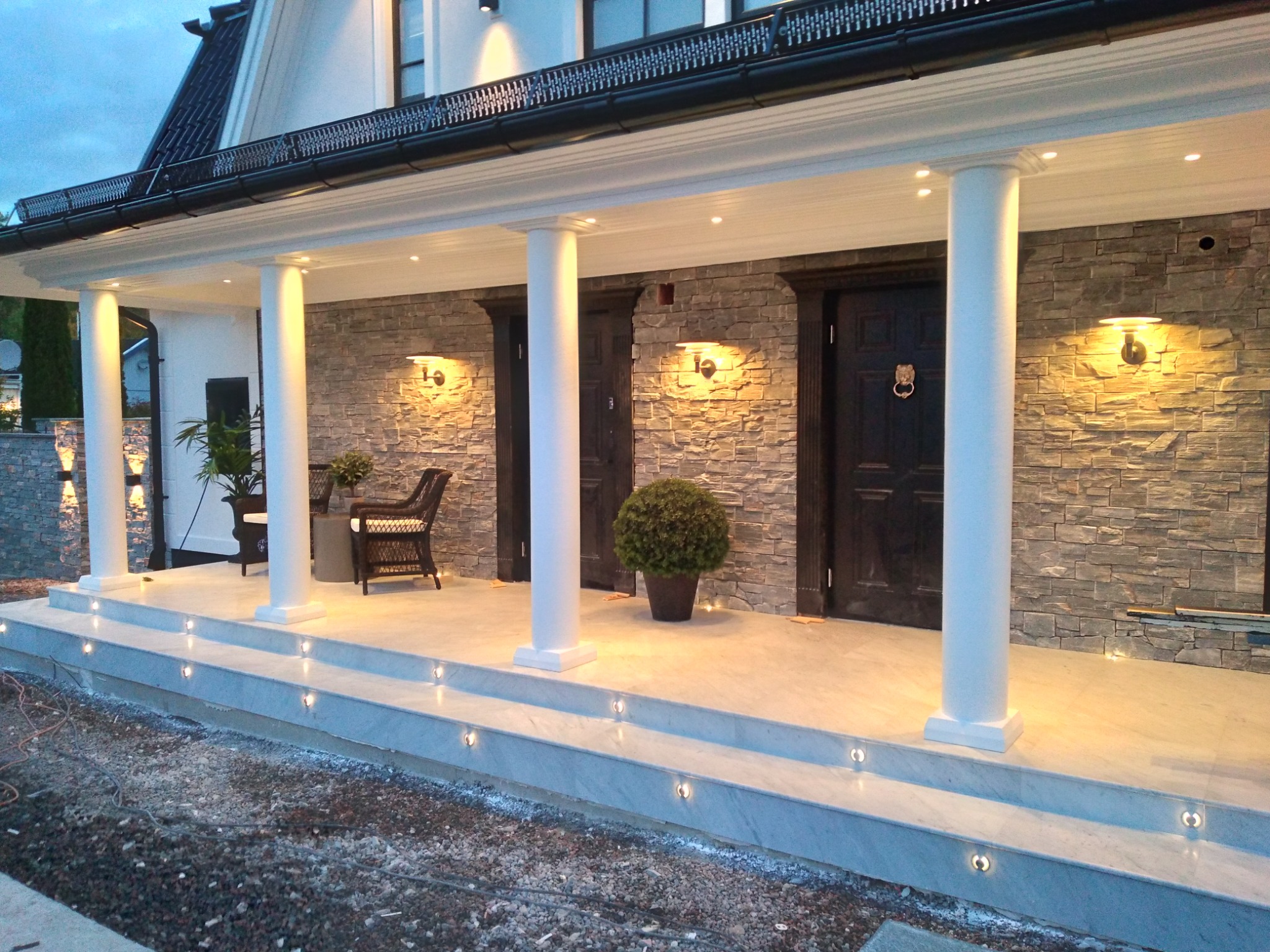 Eleganckie wejście do domu z kamienną elewacją, oświetlonymi schodami z marmuru i kolumnami, widoczne krzesło wiklinowe i rośliny w donicach.