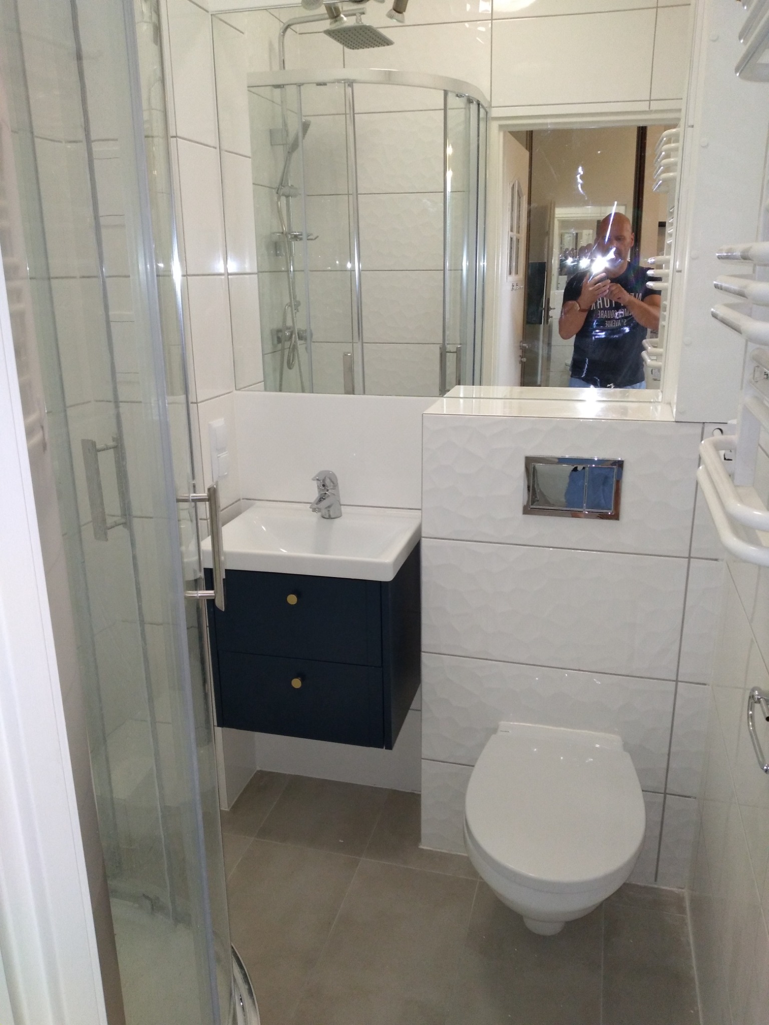 Nowoczesna łazienka po remoncie z kabiną prysznicową, umywalką z szafką w kolorze granatowym i toaletą. W lustrze odbija się osoba robiąca zdjęcie.