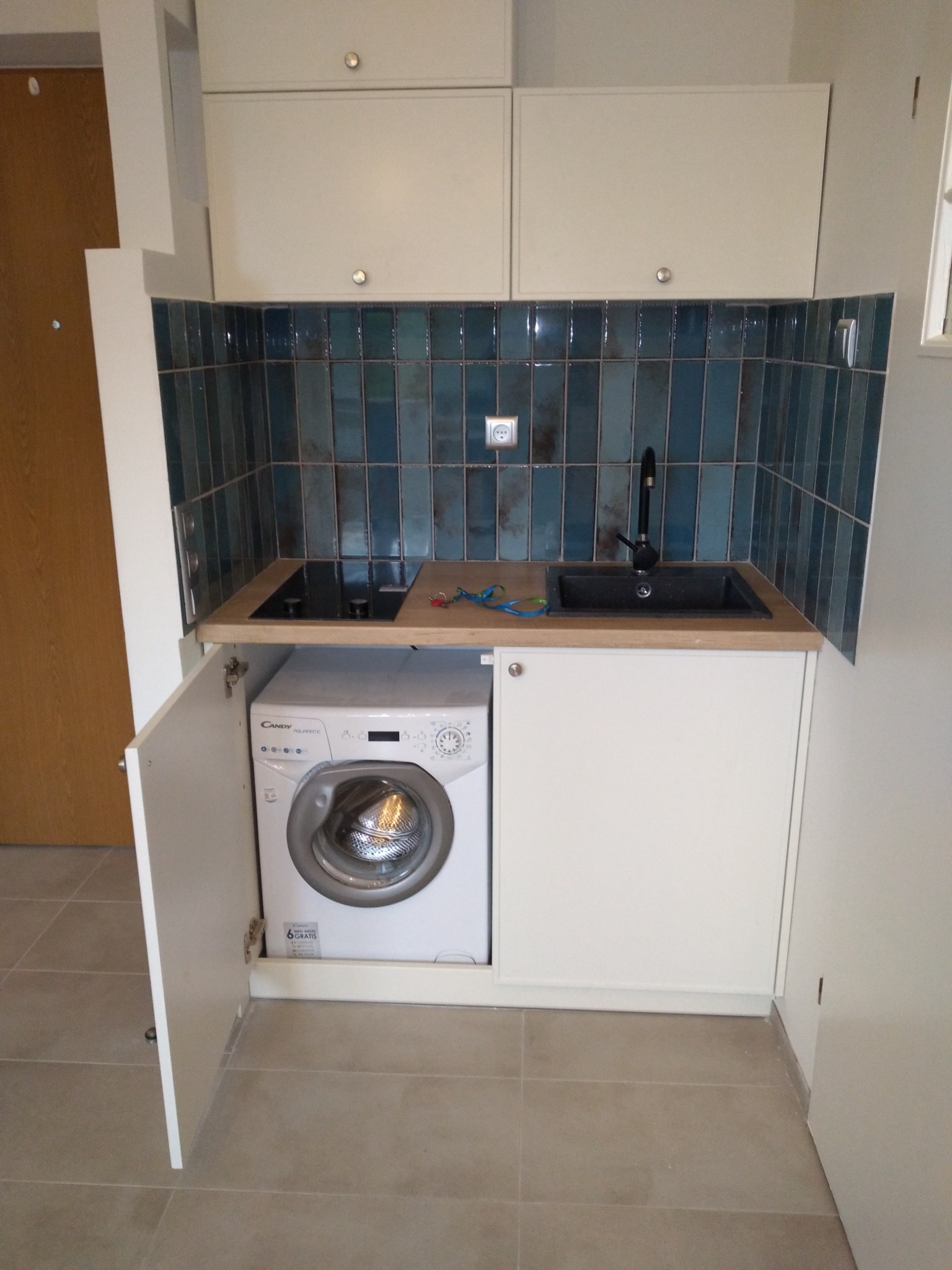 Kompaktowa zabudowa kuchenna z pralką ukrytą za frontem, niebieskie płytki na ścianie, czarny zlew i płyta indukcyjna na drewnianym blacie.