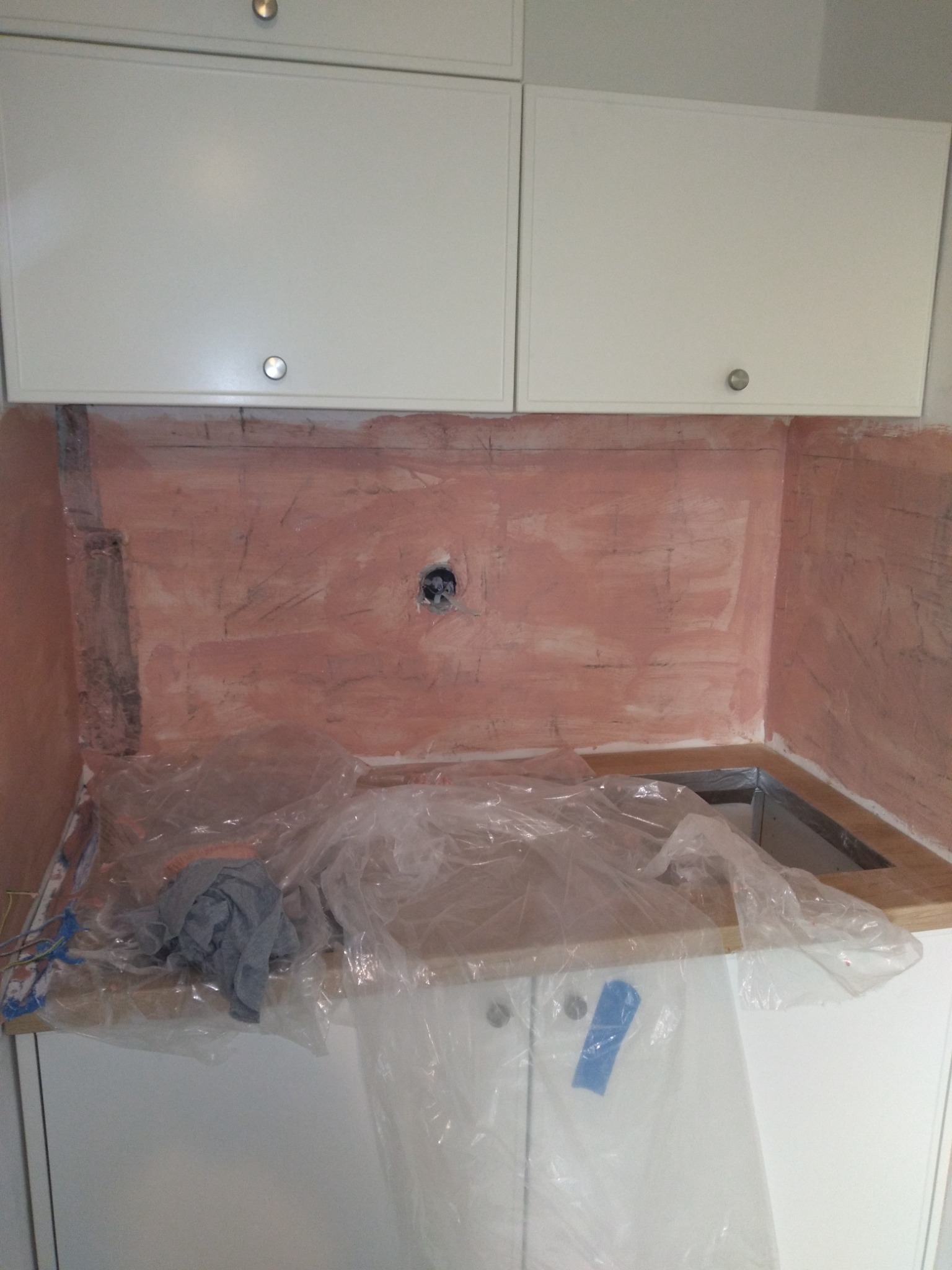 Przygotowanie ściany pod płytki w kuchni: nałożona warstwa gruntująca w kolorze różowym, widoczne ślady po ołówku, zabezpieczone folią meble kuchenne.