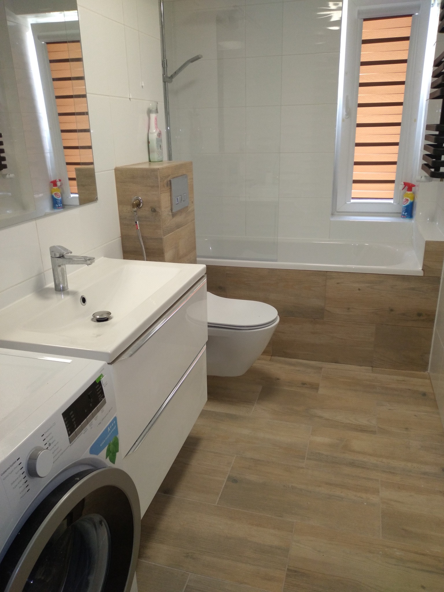 Nowoczesna łazienka wykończona w odcieniach bieli i drewna, z widoczną pralką, umywalką z szafką, toaletą podwieszaną oraz wanną obudowaną panelami imitującymi drewno. Na ścianach białe płytki,...