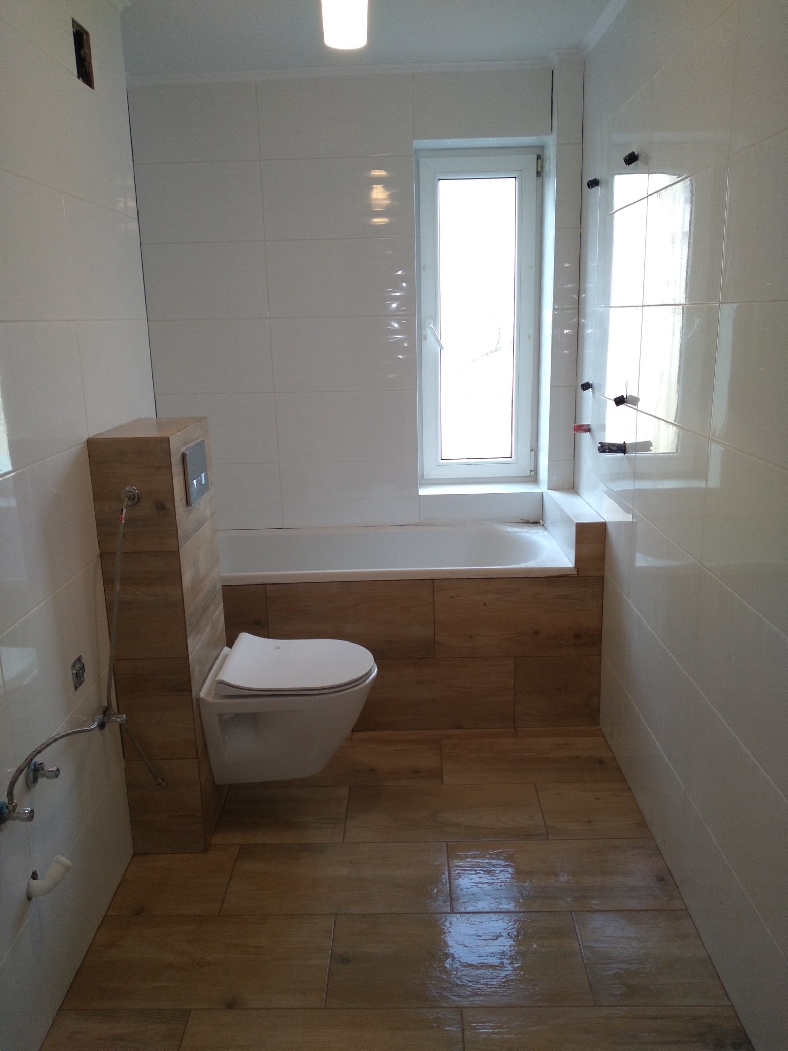 Nowoczesna łazienka z białymi płytkami ściennymi i drewnopodobnymi płytkami podłogowymi, wisząca toaleta z zabudowaną spłuczką, okno nad wanną, widoczne ślady po montażu akcesoriów.