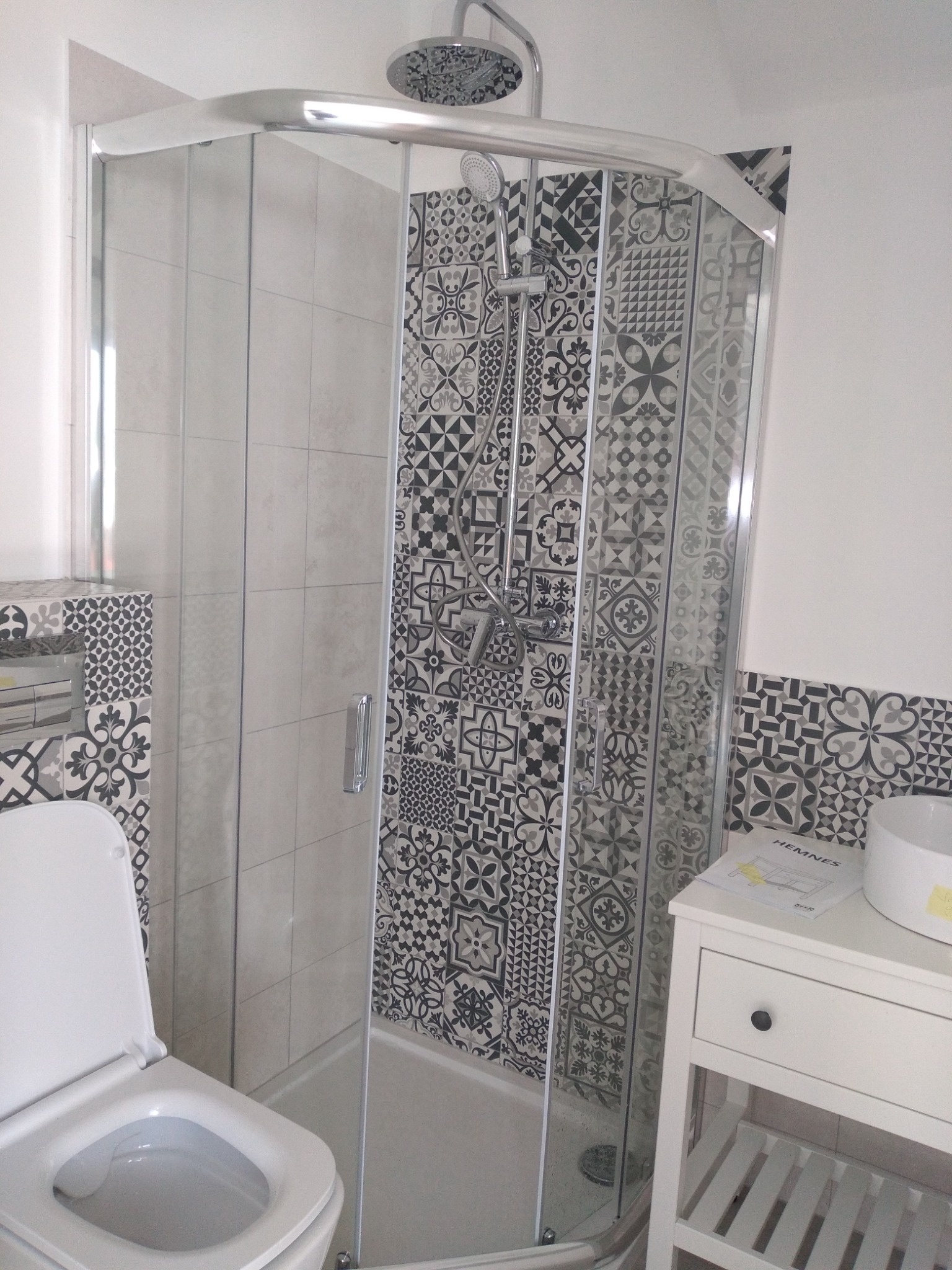 Ukończona łazienka z kabiną prysznicową z chromowanymi profilami, płytkami w geometryczne wzory, białą toaletą i umywalką na szafce Hemnes.