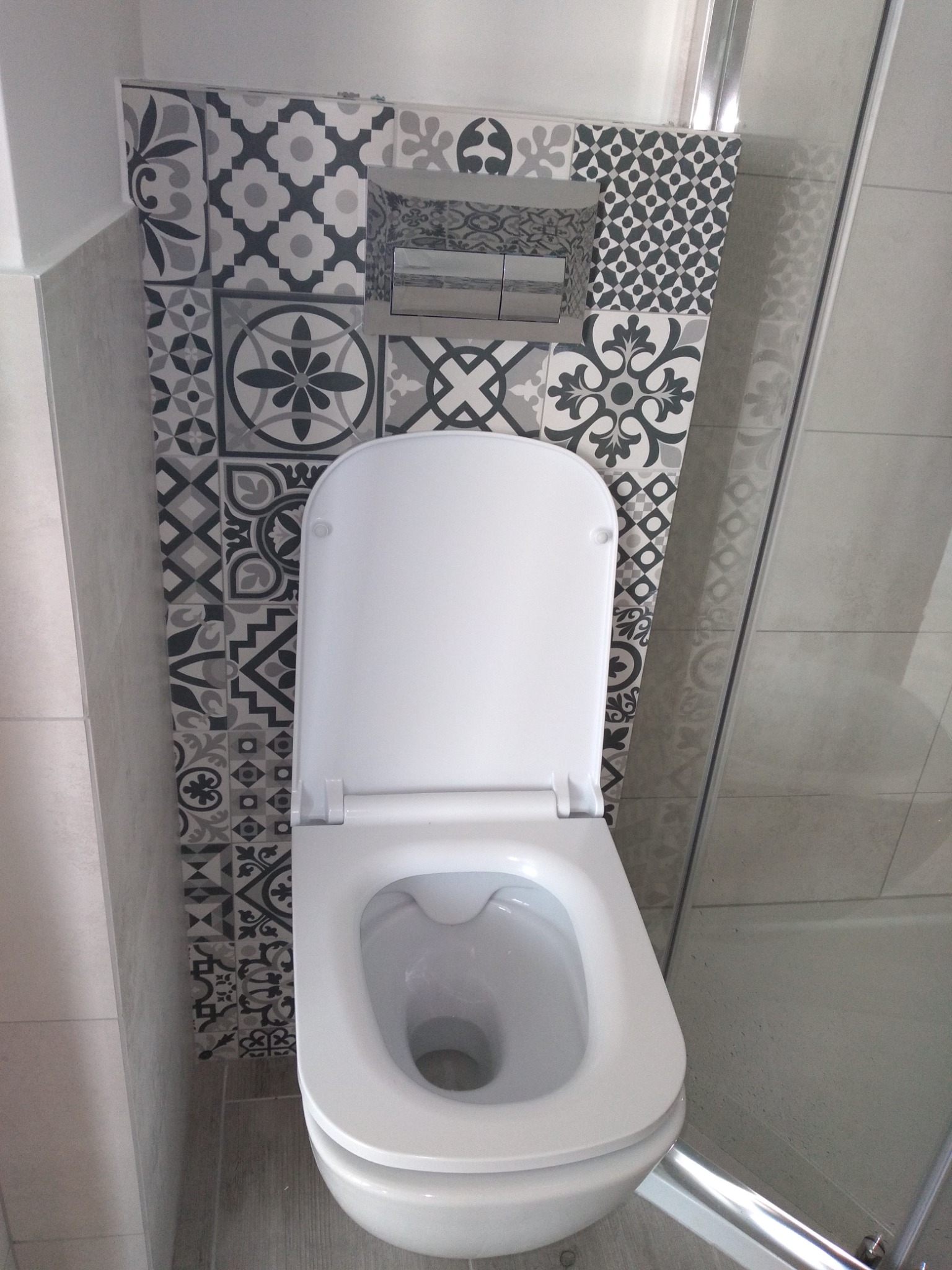 Biała toaleta z otwartą klapą, zamontowana na ścianie wyłożonej dekoracyjnymi, szaro-białymi płytkami z motywami geometrycznymi i kwiatowymi, widoczna częściowo kabina prysznicowa.