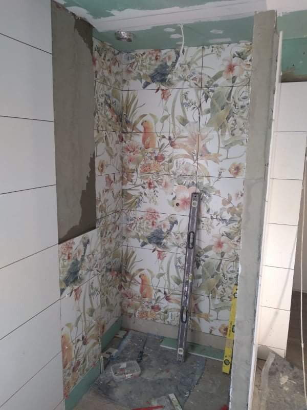 Wnętrze łazienki w trakcie układania płytek z motywem kwiatowym i papug, widoczny poziomica i narzędzia na podłodze, ściany częściowo wykończone gładkimi, białymi płytkami.