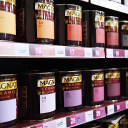 Półki w sklepie z farbami Magnat Ceramic w odcieniach beżu, pomarańczu, różu i fioletu, z widocznymi numerami i nazwami kolorów na etykietach.