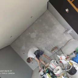 Mężczyzna w białej koszulce i ciemnych spodenkach, pochylony nad wiadrami z farbą i narzędziami malarskimi w trakcie przygotowywania ściany do malowania, widoczne ślady wcześniejszych prac...