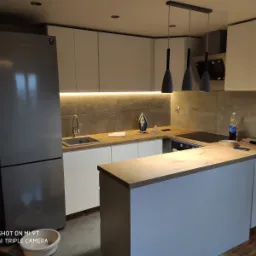 Nowoczesna kuchnia z białymi szafkami, drewnianym blatem, szarymi płytkami na ścianie i wiszącymi lampami w kształcie stożków. Na blacie stoi żelazko.