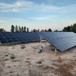 Dwa rzędy paneli fotowoltaicznych na polnej instalacji gruntowej, z widocznymi przewodami i konstrukcją wsporczą, na tle nieba z smugą kondensacyjną samolotu.