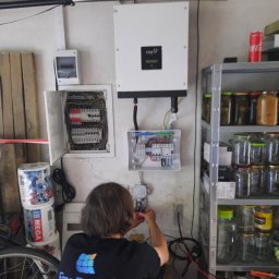 Free Energy Technology Sp. z o.o. - Instalacja elektryczna paneli słonecznych we Wrocławiu: elektryk w koszulce Free Energy Technology łączy przewody w skrzynce rozdzielczej obok inwertera FOX ESS. W tle regał z przetworami i puszka...
