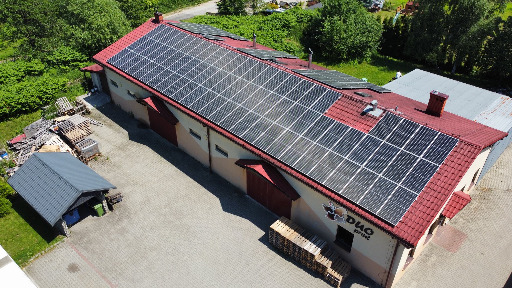 Widok z góry na budynek z czerwonym dachem pokrytym panelami słonecznymi; widoczne palety i budynek gospodarczy obok.