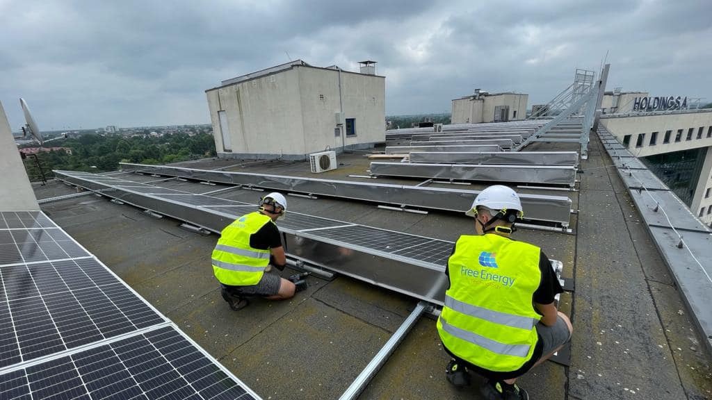 Montaż paneli słonecznych na dachu budynku przez dwóch pracowników w kaskach i kamizelkach odblaskowych z logo 'Free Energy'. Widoczny fragment miasta w tle.