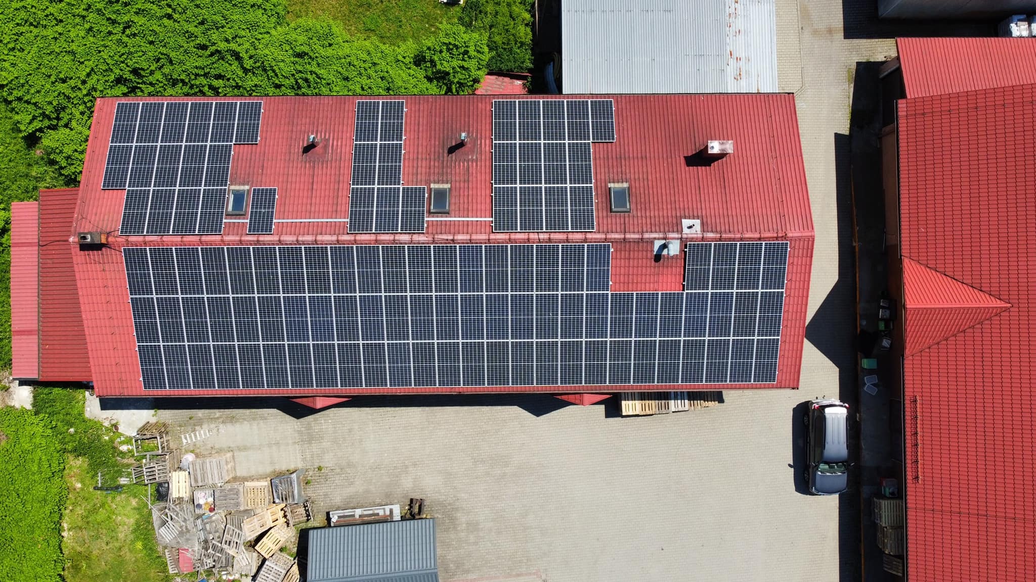 Widok z lotu ptaka na czerwony dach budynku przemysłowego pokryty panelami słonecznymi, z widocznymi świetlikami, paletami i samochodem na parkingu.
