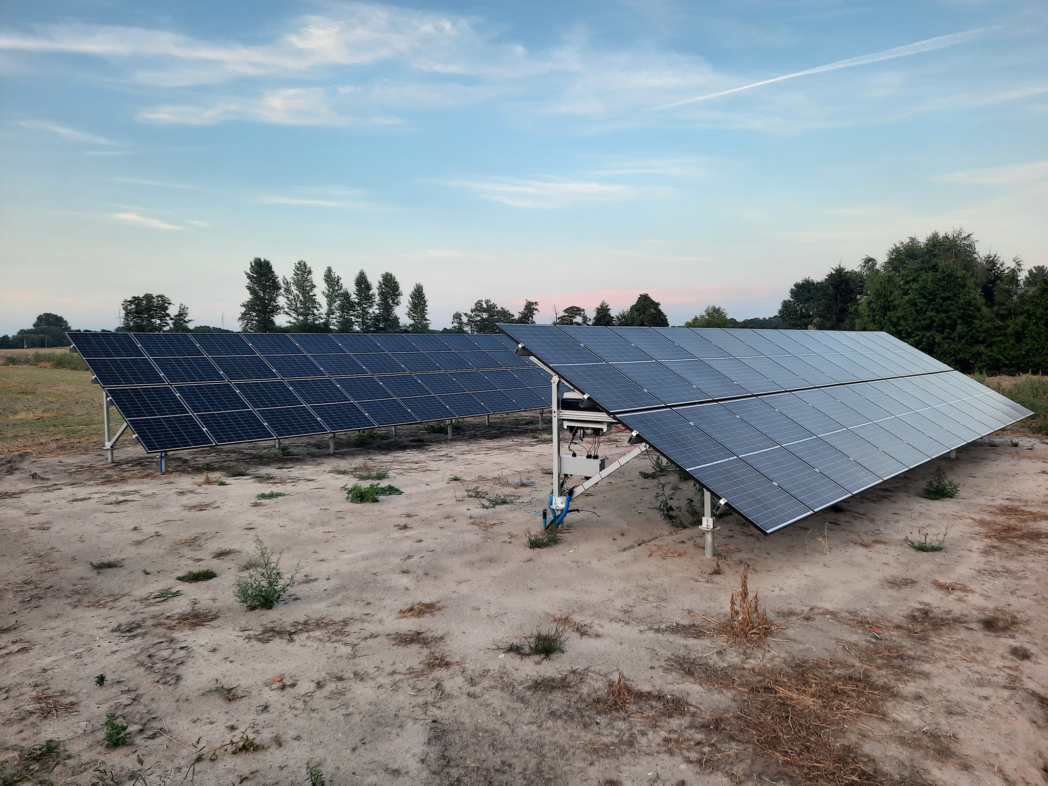 Dwa rzędy paneli fotowoltaicznych na polnej instalacji gruntowej, z widocznymi przewodami i konstrukcją wsporczą, na tle nieba z smugą kondensacyjną samolotu.