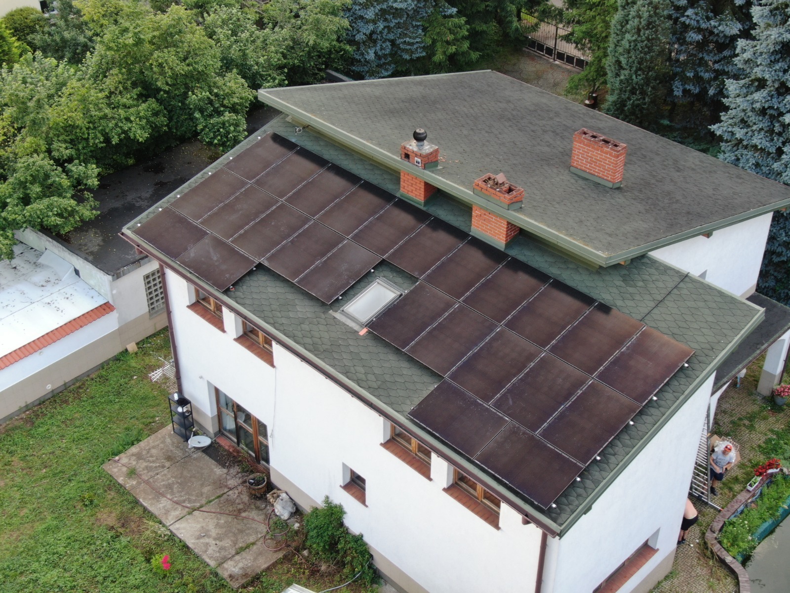 Widok z góry na biały dom z szarym dachem pokrytym panelami słonecznymi; widoczny komin i okno dachowe.