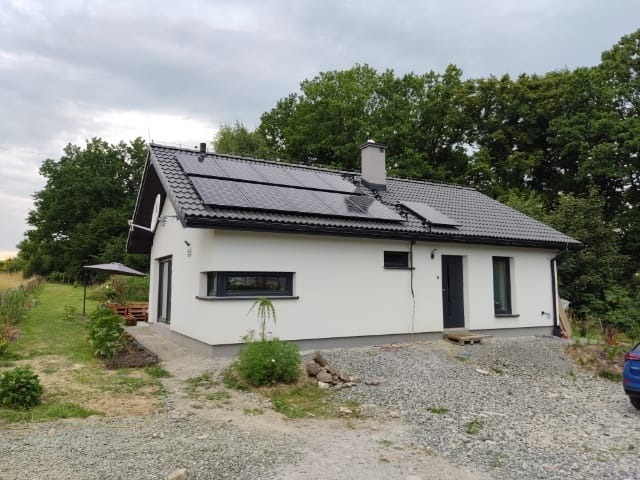 Biały, parterowy dom z panelami słonecznymi na ciemnym dachu, widziany z perspektywy ogrodu z widocznym fragmentem niebieskiego samochodu po prawej stronie.