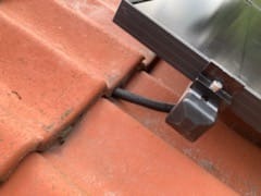 Szczegół montażu kabla fotowoltaicznego na czerwonej dachówce, widoczny element mocujący i fragment panelu.