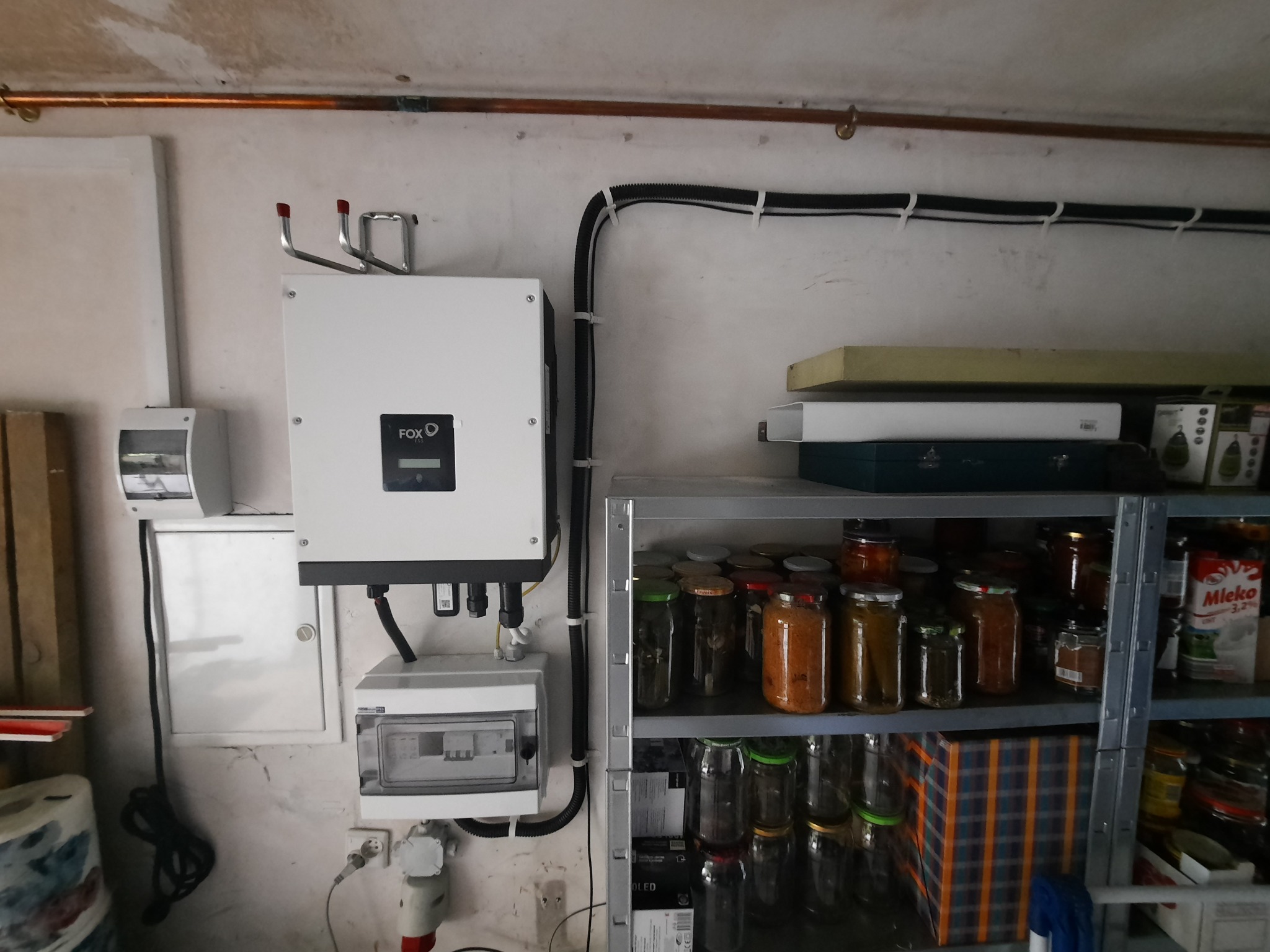 Biały inwerter marki FOX podłączony do skrzynki elektrycznej, zamontowany na ścianie w pomieszczeniu gospodarczym, obok metalowego regału z przetworami w słoikach.