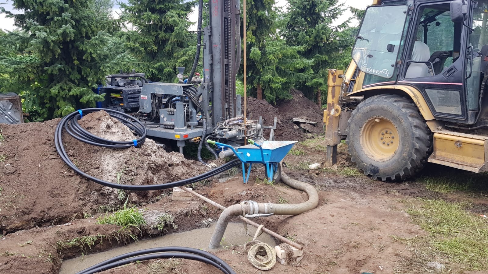 Wiertnica pionowa i koparko-ładowarka na placu budowy, przygotowanie do instalacji gruntowej pompy ciepła, widoczne rury kolektora gruntowego i niebieska taczka.