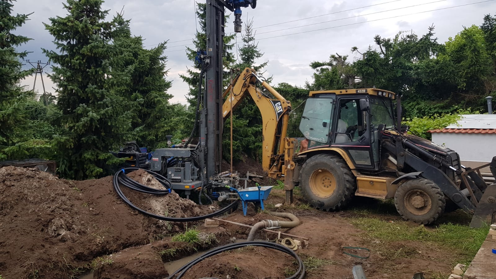 Wiertnica hydrauliczna i koparko-ładowarka CAT 432D na placu budowy, prawdopodobnie podczas wiercenia otworów pod instalację pompy ciepła lub studni głębinowej.