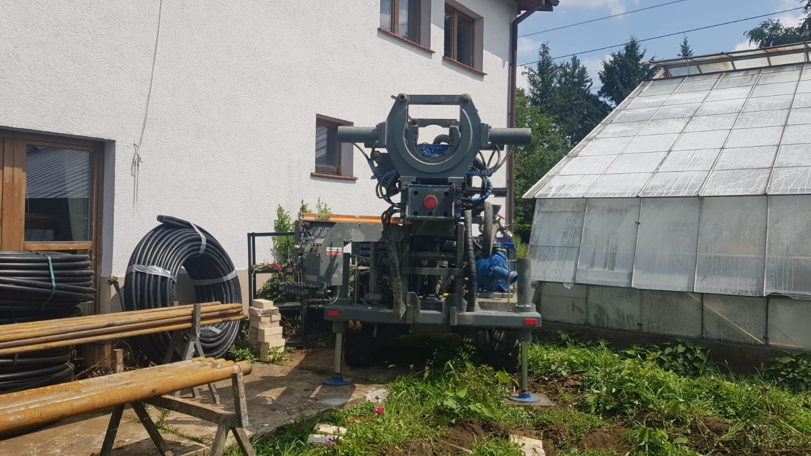 Wiertnica geotermalna ustawiona na trawniku obok domu i szklarni, widoczne rury do instalacji podziemnej.
