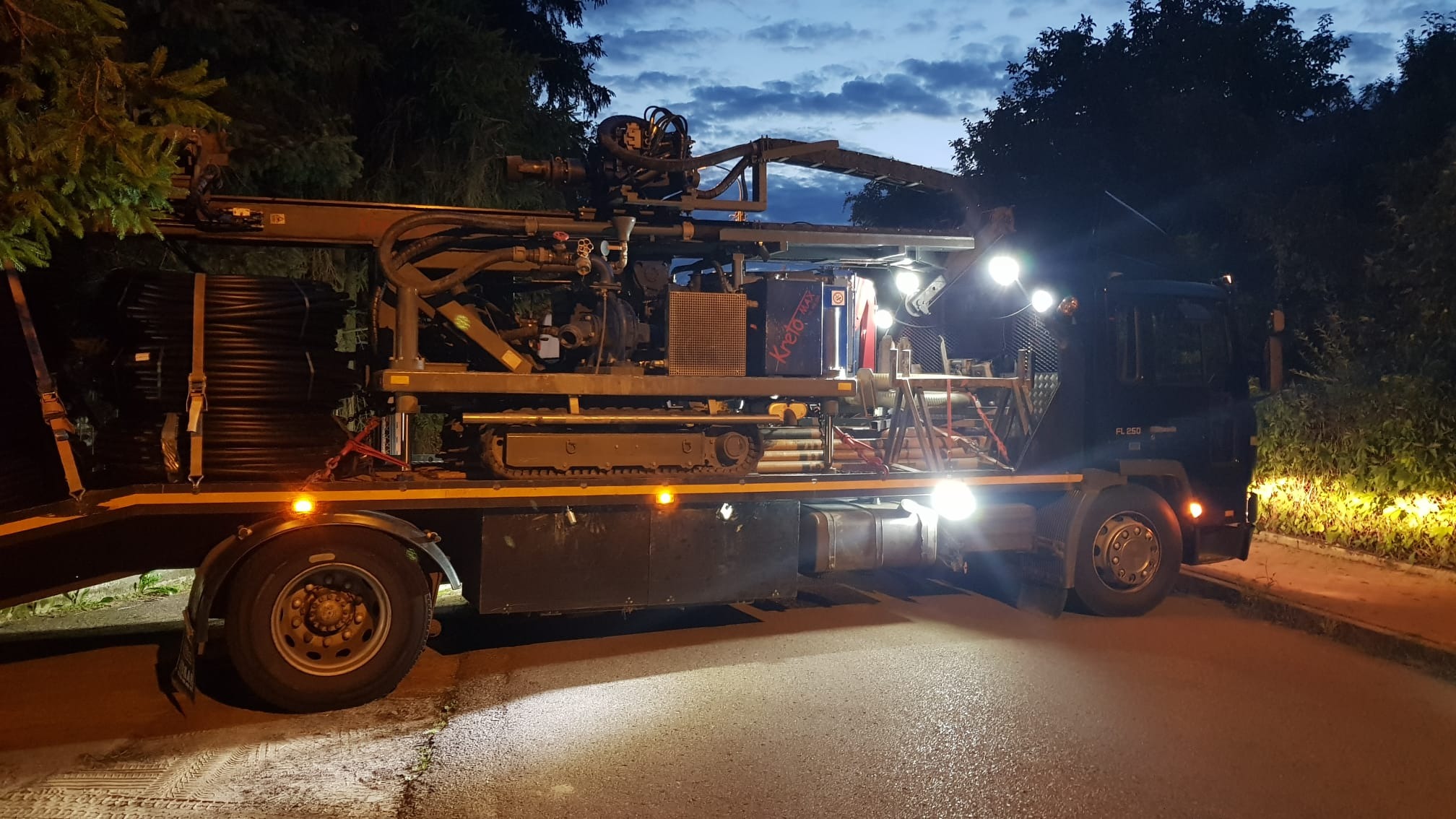 Ciężarówka z wiertnicą do odwiertów geotermalnych, oświetlona reflektorami, na tle wieczornego nieba i drzew.