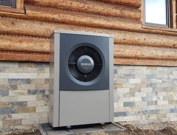 Zewnętrzna jednostka pompy ciepła Buderus zamontowana przy drewnianym domu z kamienną podmurówką, widoczna instalacja na betonowej podstawie.