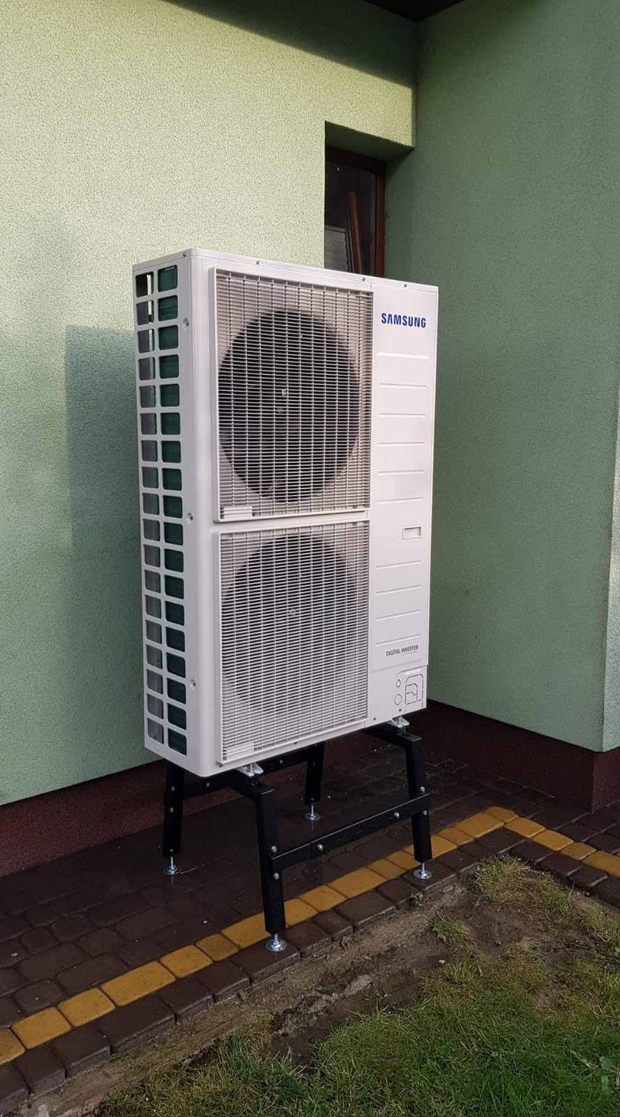 Zewnętrzna jednostka pompy ciepła Samsung Digital Inverter, zamontowana na czarnym stelażu z regulowanymi nóżkami, stoi na kostce brukowej przy ścianie budynku o oliwkowym odcieniu.