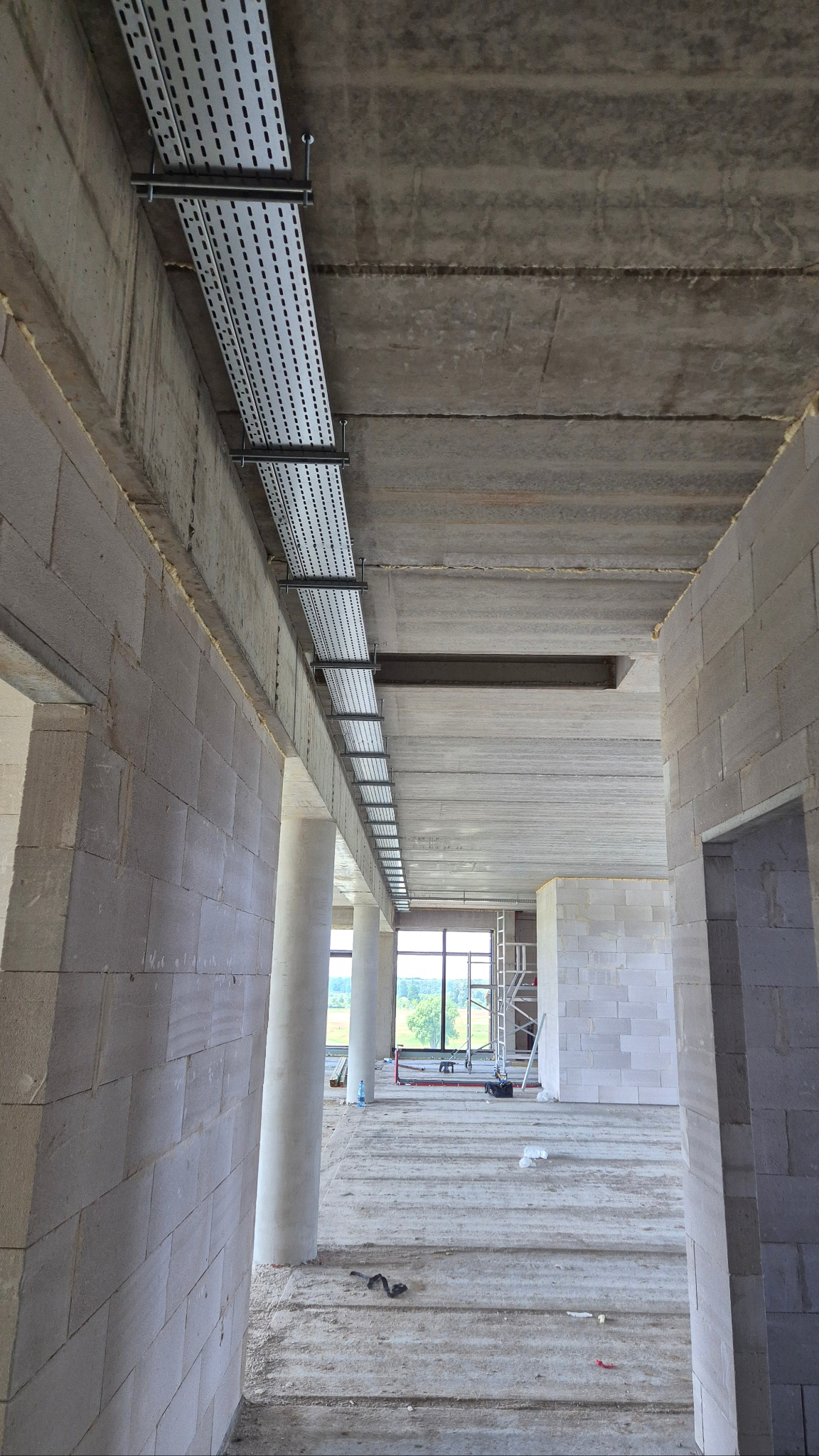 Metalowe korytka kablowe zamontowane pod betonowym stropem w surowym wnętrzu budynku. Widoczne mocowania i perforowana konstrukcja korytek. Surowy stan wykończenia.