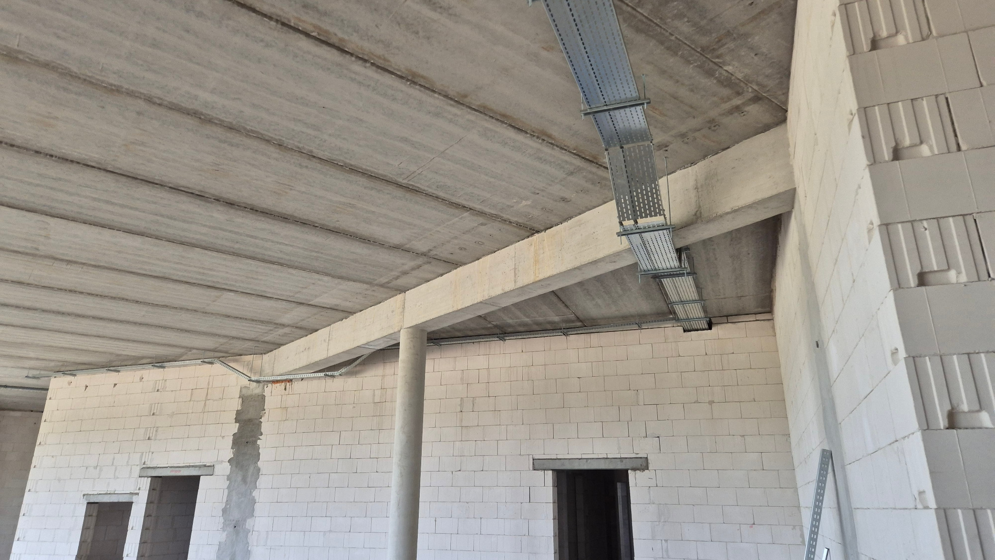 Surowy stan budynku z widocznymi korytkami kablowymi pod sufitem. Widoczne betonowe stropy i ściany z białych bloczków. Koryta zamontowane równolegle do belek stropowych.