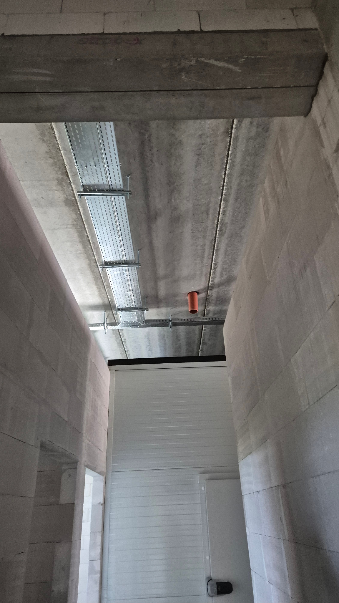 Metalowe korytka kablowe zamontowane pod betonowym sufitem w surowym wnętrzu. Widoczny fragment instalacji i pomarańczowa rura. Ściany z betonu komórkowego.