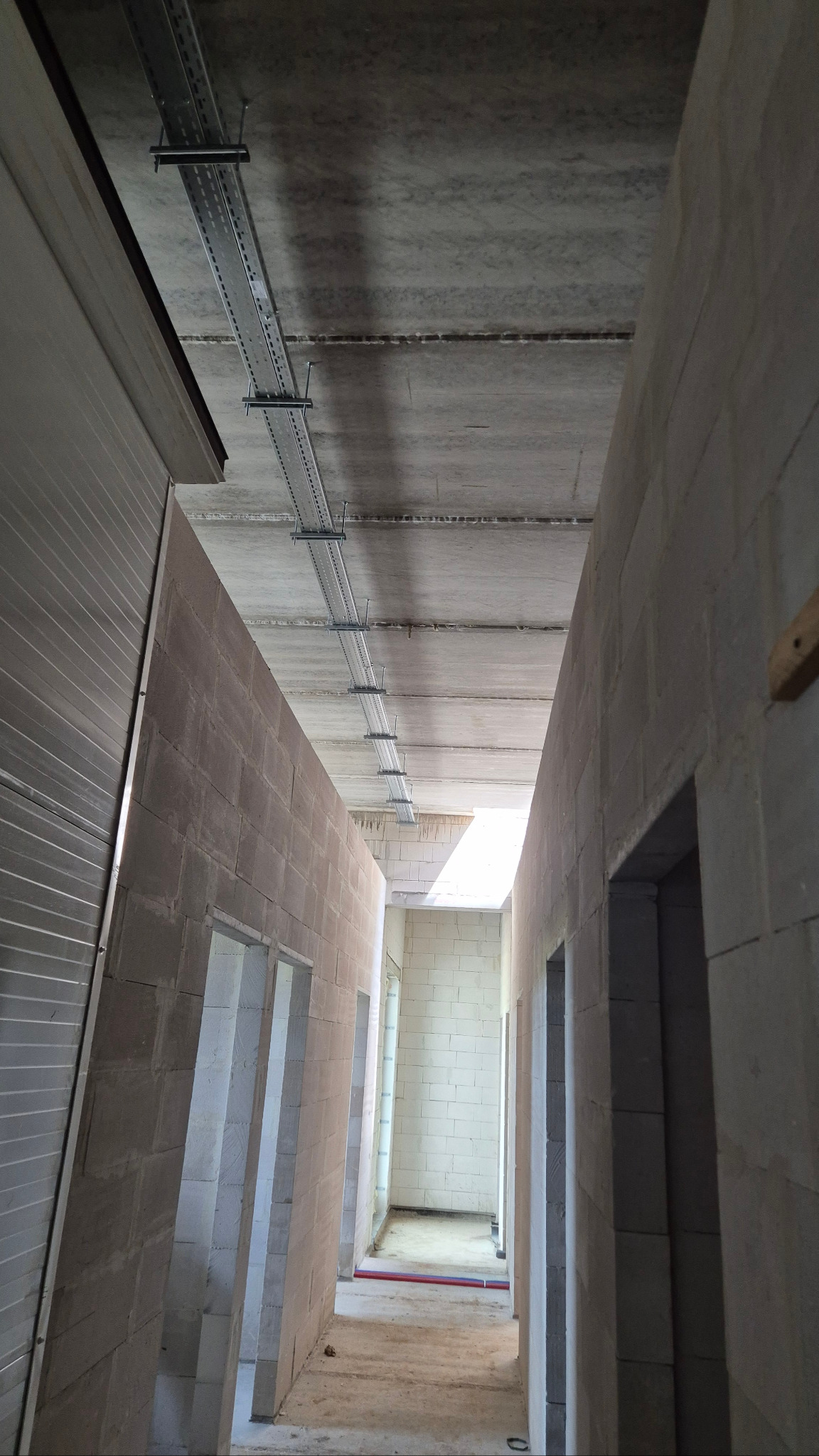 Metalowe korytka kablowe zamontowane pod betonowym sufitem w surowym korytarzu budynku w trakcie budowy. Widoczne ściany z pustaków i otwory drzwiowe.