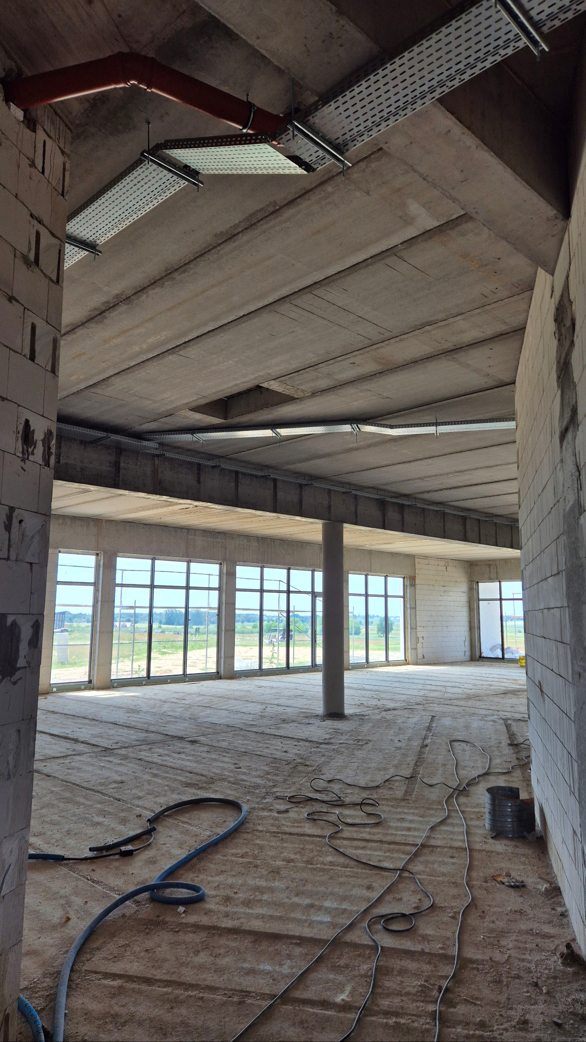 Wnętrze surowego biura z widocznymi instalacjami pod sufitem, kablami na podłodze i dużymi oknami z widokiem na zewnątrz. Stan deweloperski.