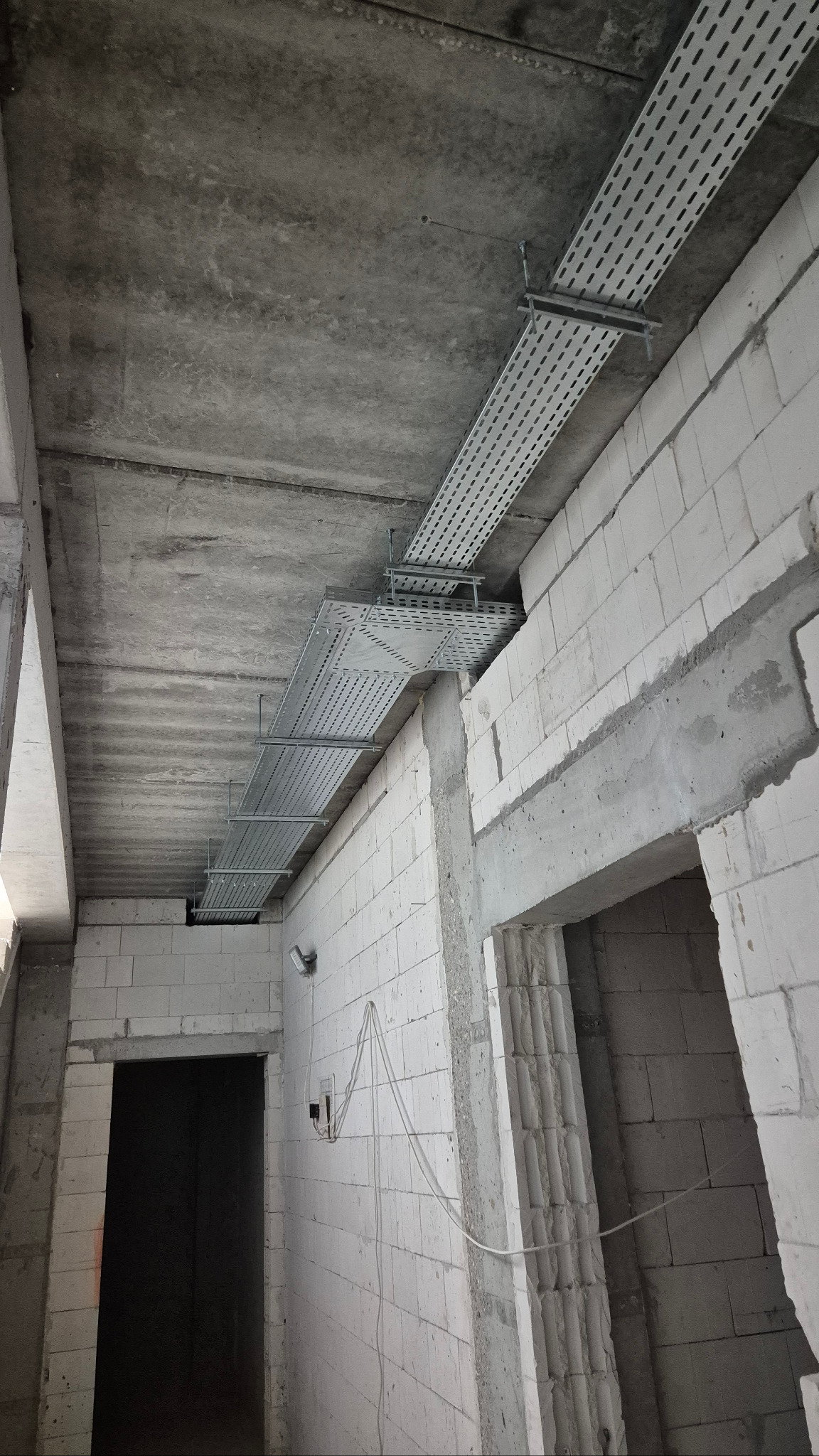 Szare koryta kablowe pod betonowym sufitem w surowym wnętrzu. Widoczne białe ściany z bloków i otwory drzwiowe. Instalacja w trakcie realizacji, surowy stan.