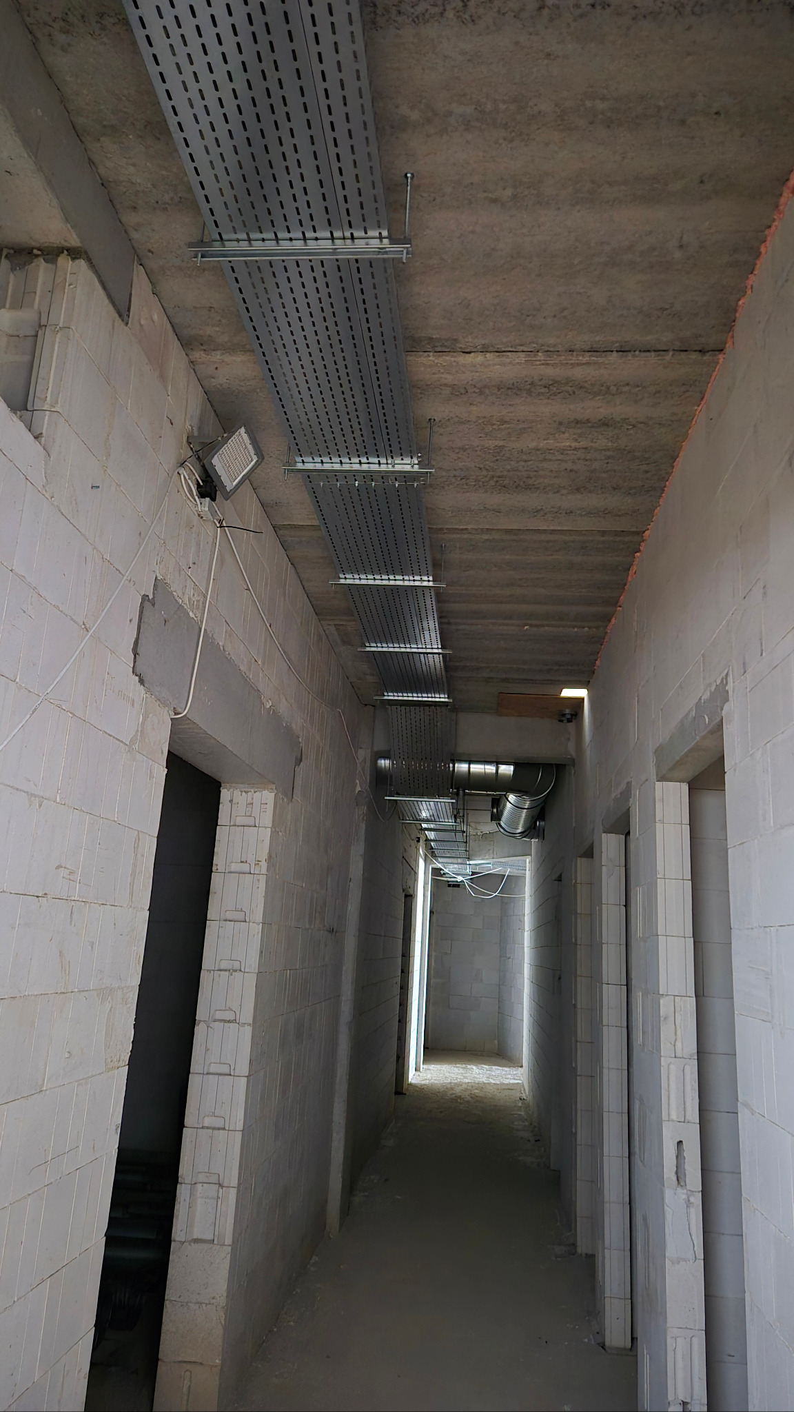 Korytarz w surowym stanie z zamontowanymi metalowymi korytkami kablowymi pod sufitem, widoczne przewody i lampa. Ściany z białych bloczków.