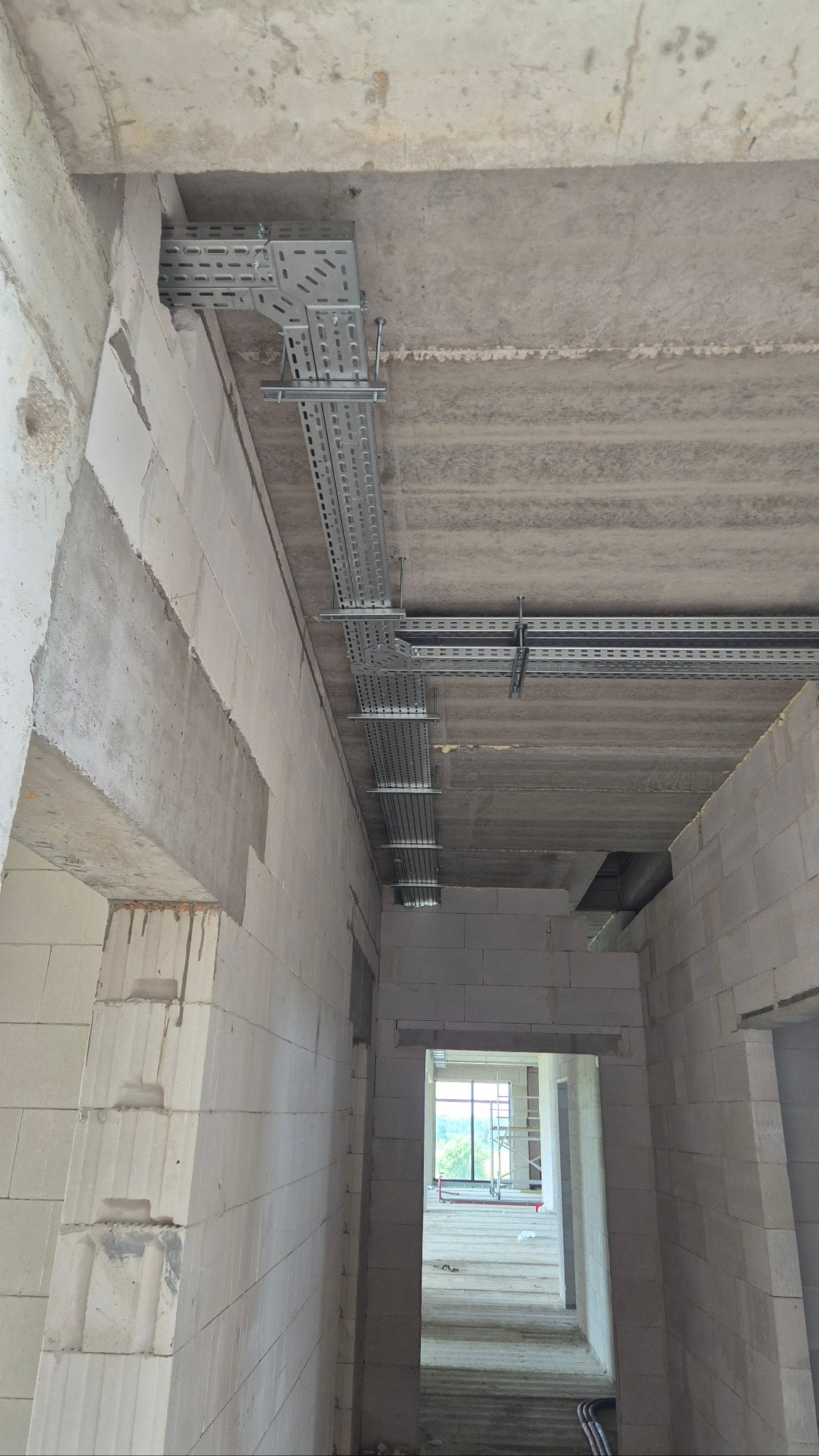 Szara, perforowana trasa kablowa zamontowana pod betonowym stropem w surowym wnętrzu budynku z białych bloczków. Widok z dołu, perspektywa korytarza.