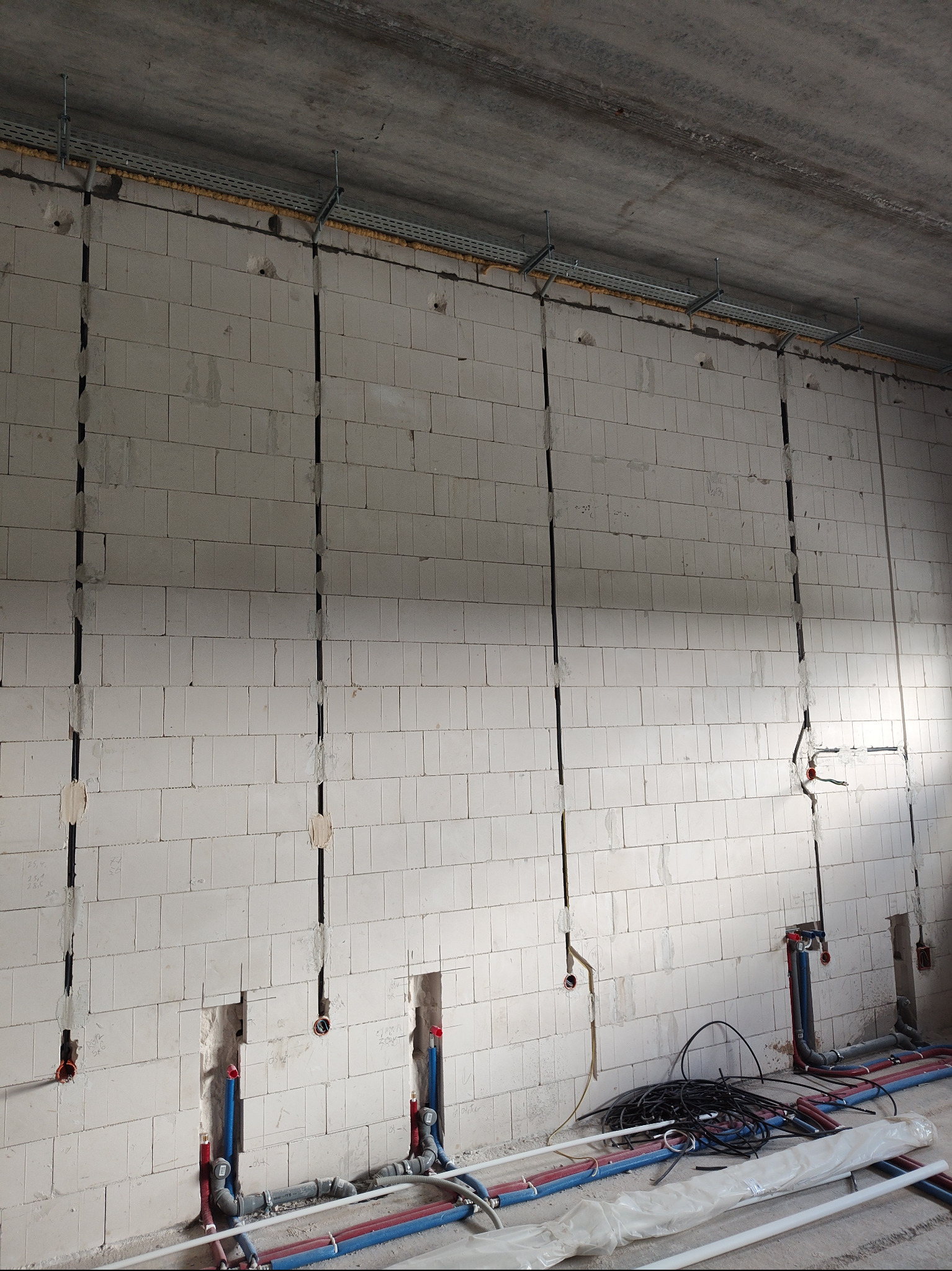 Surowa ściana z betonu komórkowego z pionowymi bruzdami na przewody elektryczne i rury. Widoczne kable, rury PEX i metalowe korytka kablowe pod sufitem.