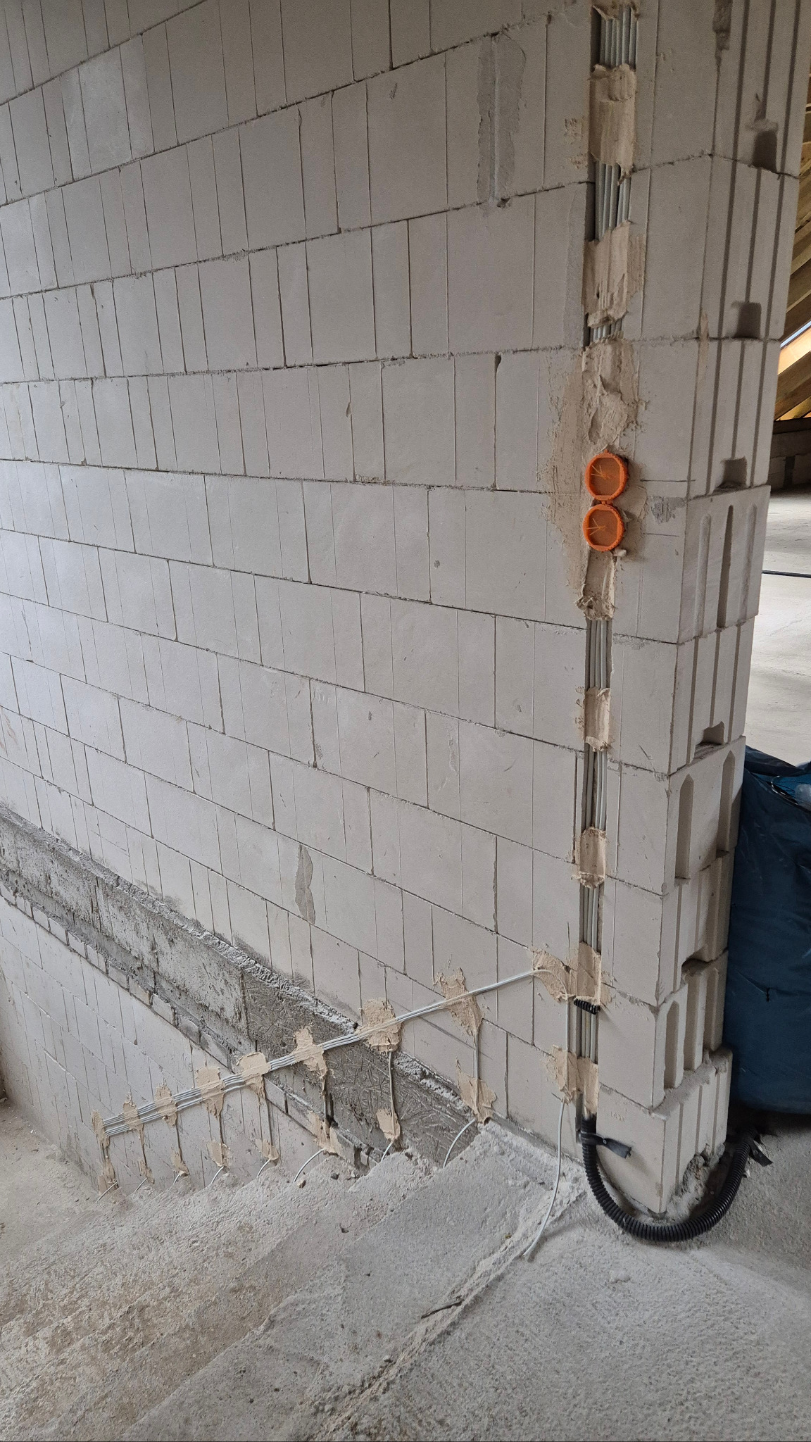 Ściana z betonu komórkowego z bruzdami, w których poprowadzono metalowe rurki instalacji elektrycznej. Widoczne pomarańczowe puszki pod gniazdka i kable. Etap prac instalacyjnych.