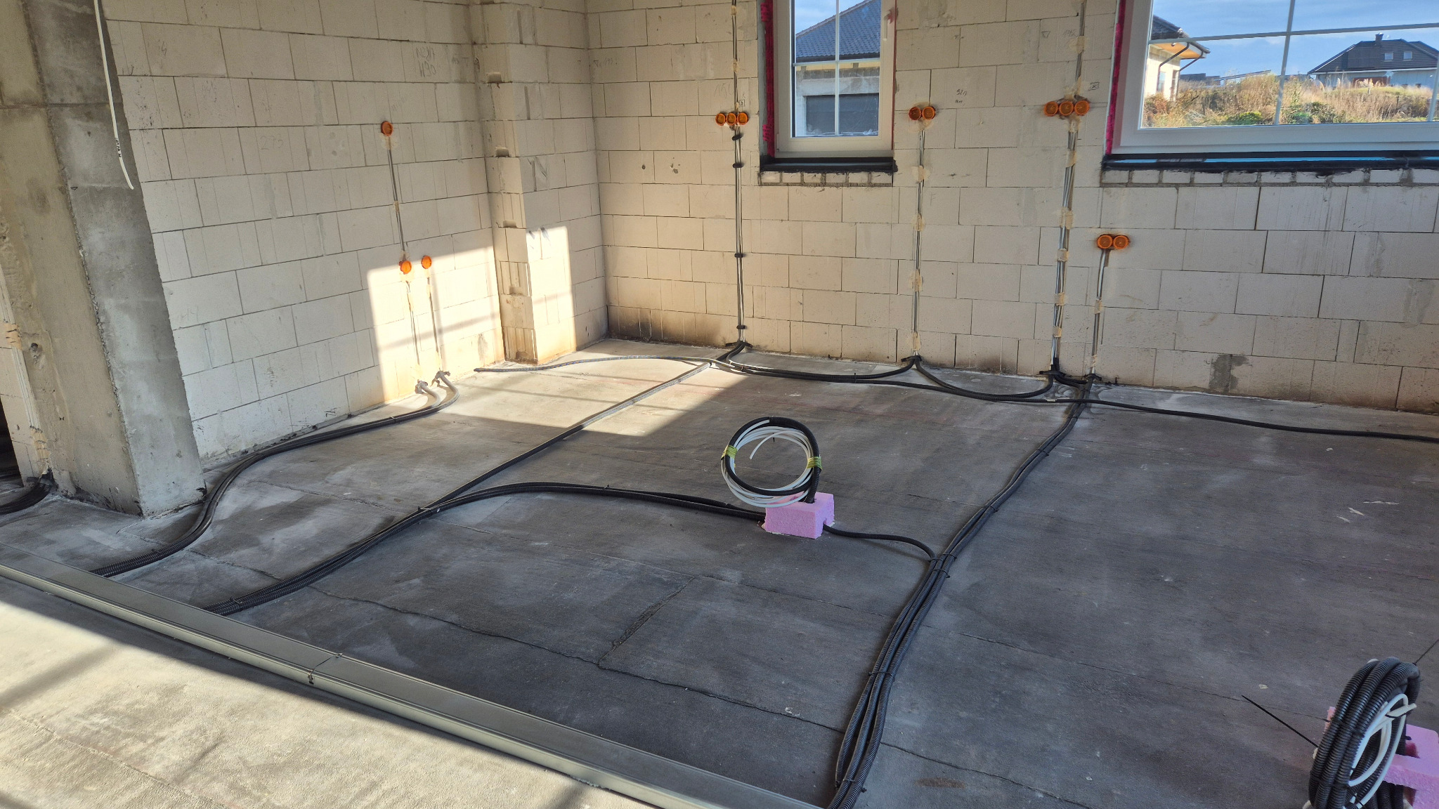 Surowy stan: przewody elektryczne ułożone na podłodze wewnątrz budynku z białego pustaka. Widoczne puszki instalacyjne na ścianach i okna z widokiem na domy.