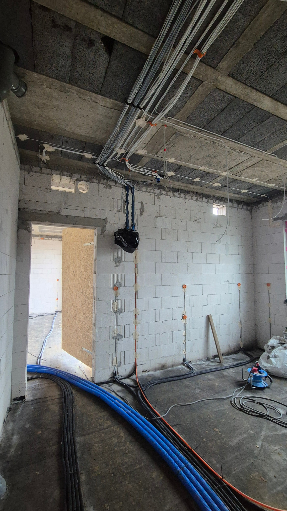 Surowy stan: instalacja elektryczna w budynku z białych bloczków. Kable w peszlach prowadzone po suficie i ścianach, widoczne puszki. Podłoga wylewka.