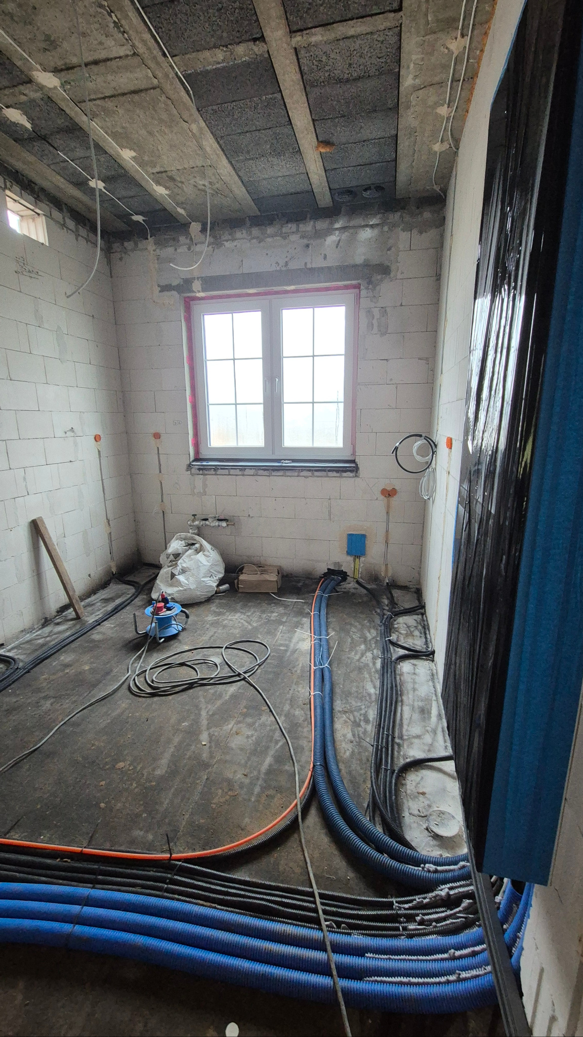 Wnętrze w trakcie prac instalacyjnych: widoczne kable elektryczne w peszlach (niebieskie i czarne) ułożone na podłodze, ściany z białego bloczka, okno w tle.