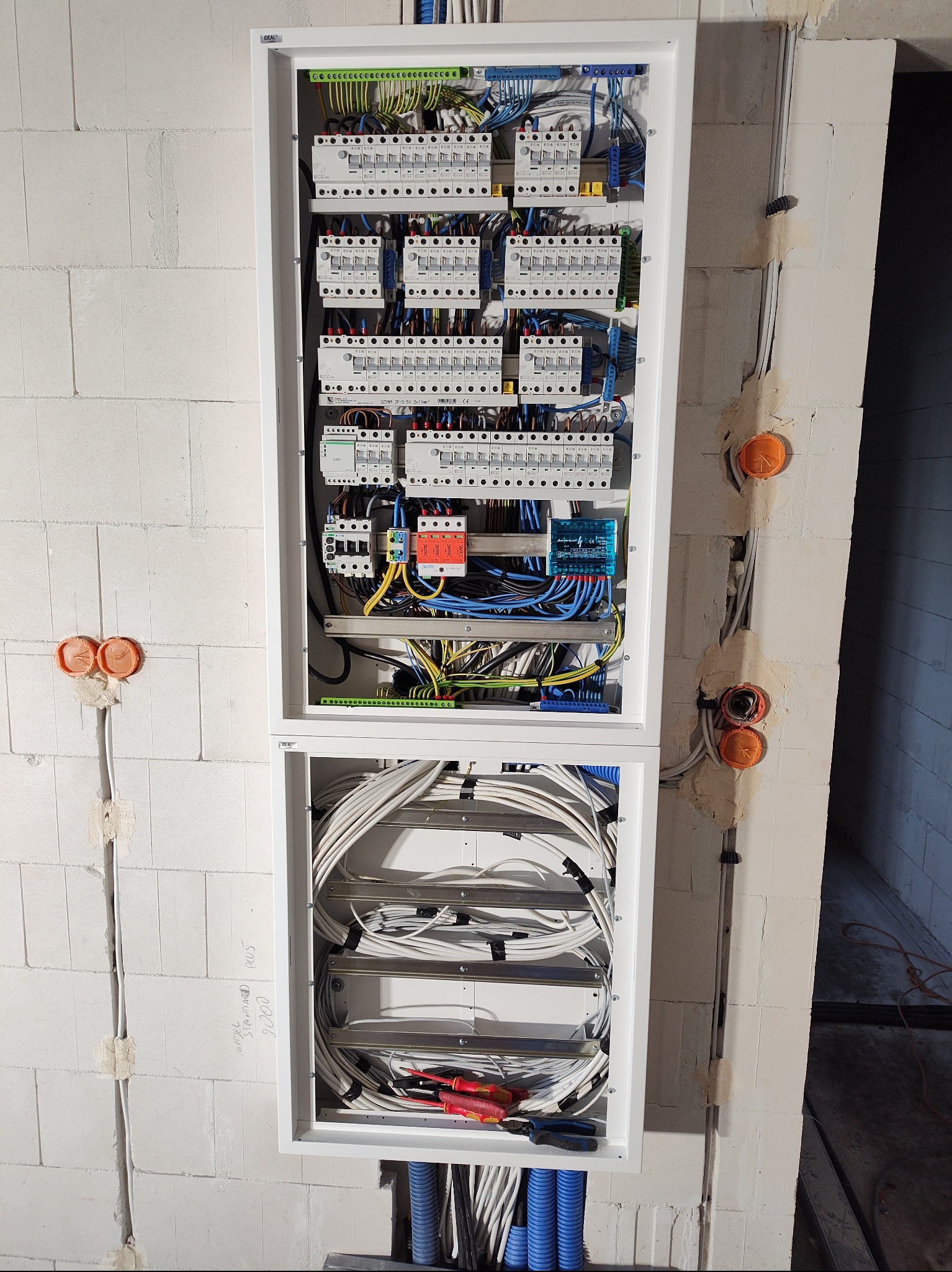 Rozdzielnia elektryczna w białej obudowie, widoczne zabezpieczenia, przewody i narzędzia. Instalacja w surowym wnętrzu z bloczków.