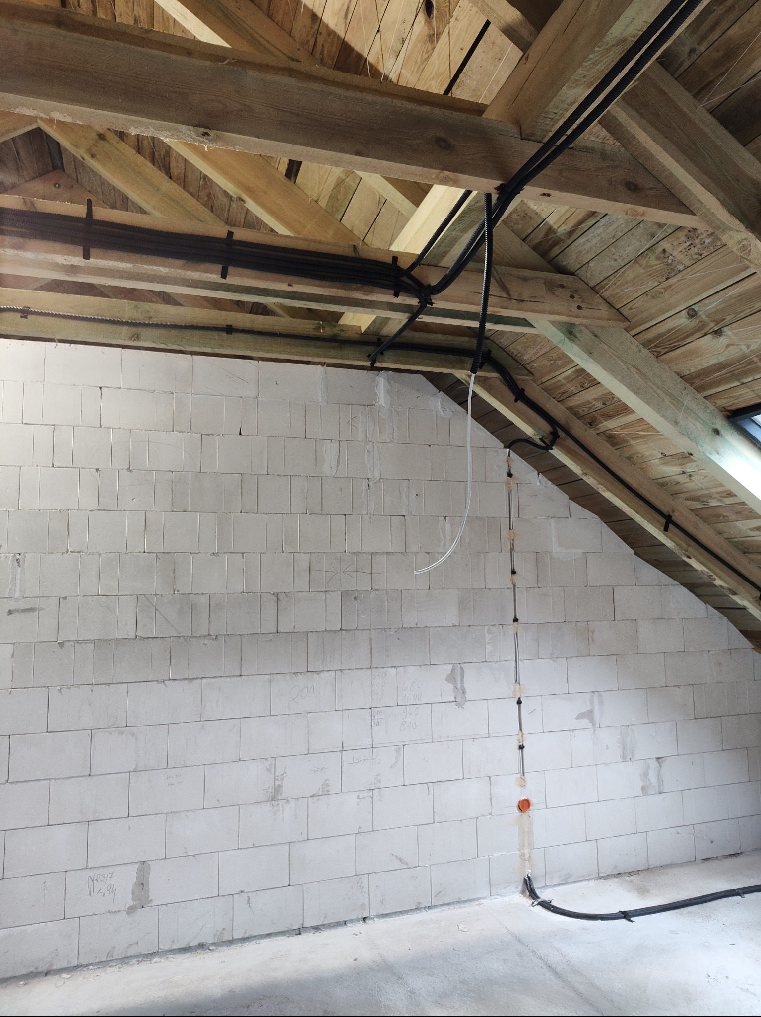 Instalacja przewodów elektrycznych w surowym wnętrzu poddasza z drewnianą konstrukcją stropu i ścianami z białych bloczków. Widoczne kable i puszki instalacyjne.