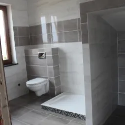 Wykończona łazienka w szaro-białych płytkach z widocznym sedesem podwieszanym i brodzikiem z mozaikowym wykończeniem. Widoczne płytki imitujące marmur na ścianach.