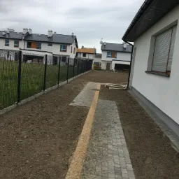 Przygotowany teren pod trawnik z ułożonym fragmentem chodnika z kostki brukowej, widoczne rury nawadniające, w tle zabudowa szeregowa.