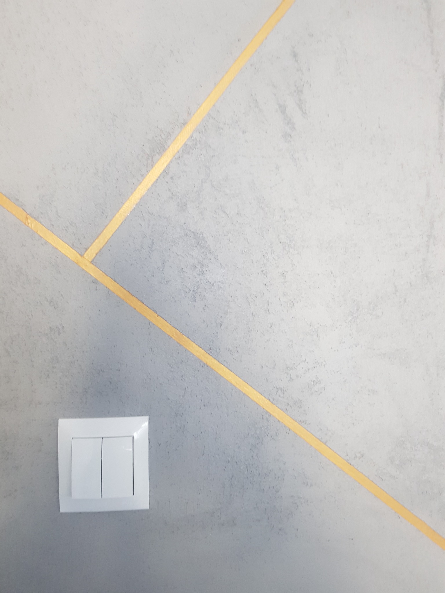 Szara ściana z dekoracyjnymi, złotymi paskami i białym podwójnym włącznikiem światła.