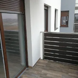 Zewnętrzny widok balkonu z brązowymi roletami opuszczonymi na oknach, drewnopodobna podłoga z płytek oraz nowoczesne balustrady balkonowe z poziomych listew.